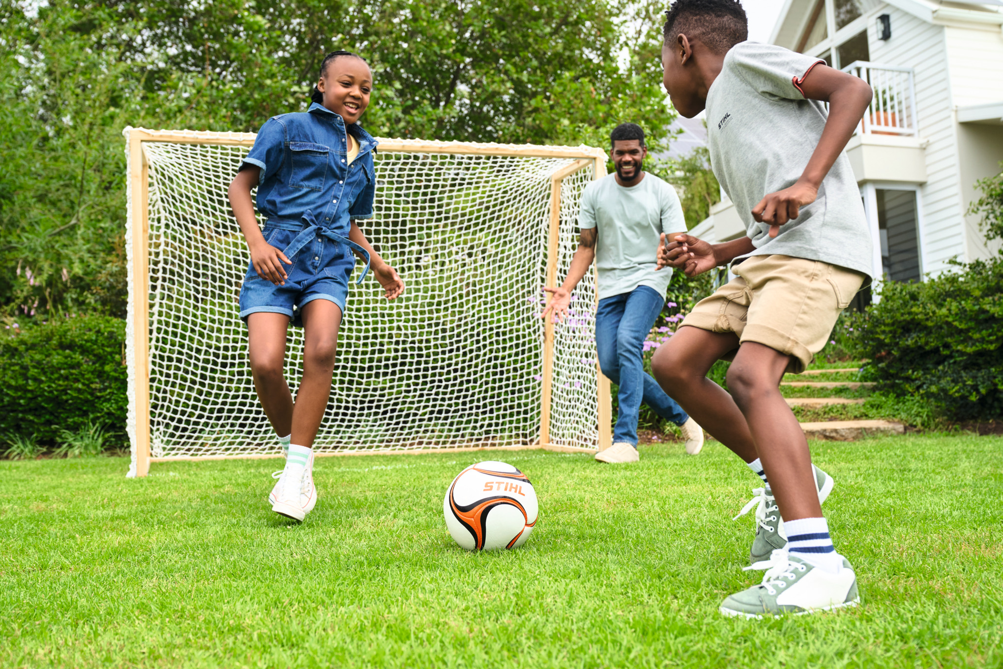 Três crianças jogam futebol num espaço verde com uma goleira de futebol em madeira