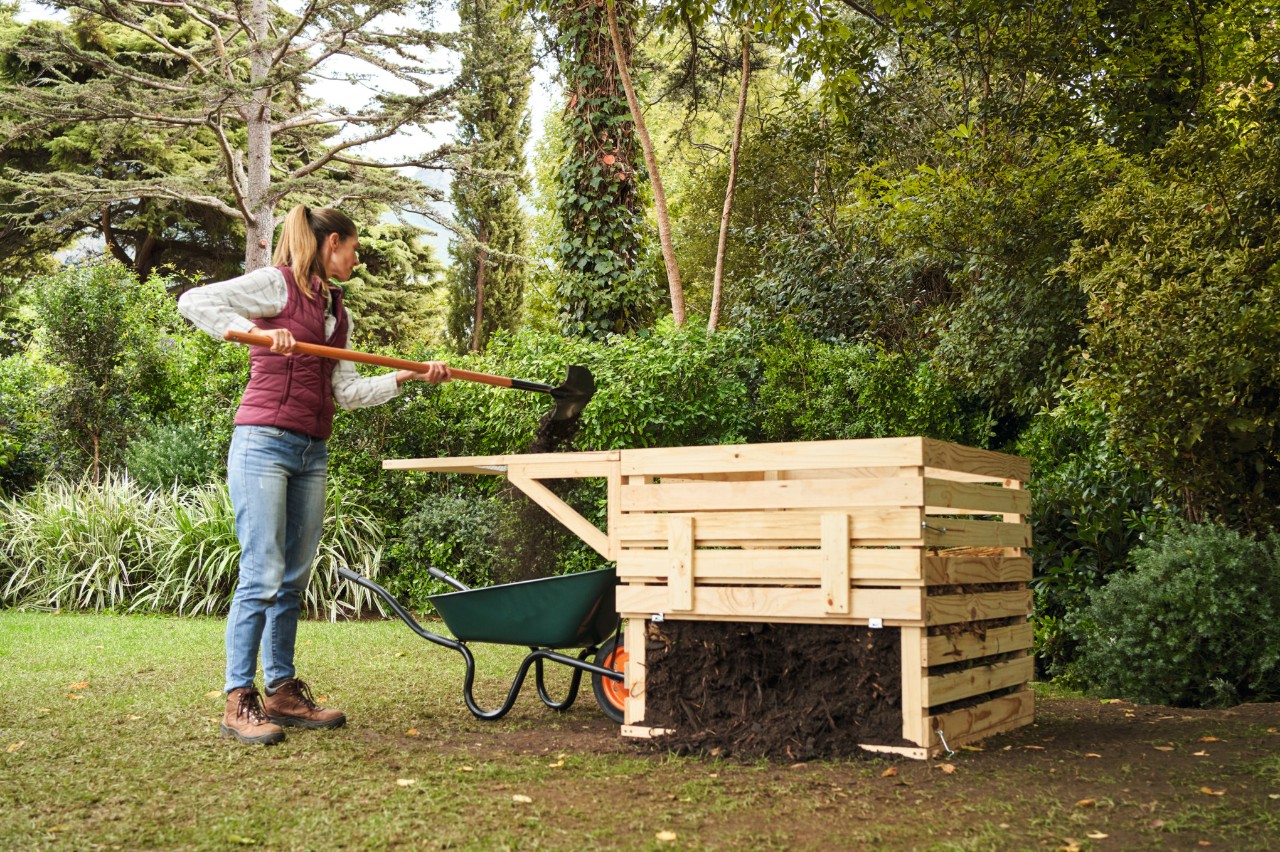 Uma mulher num canto do jardim a peneirar composto em bruto junto a composteira de madeira