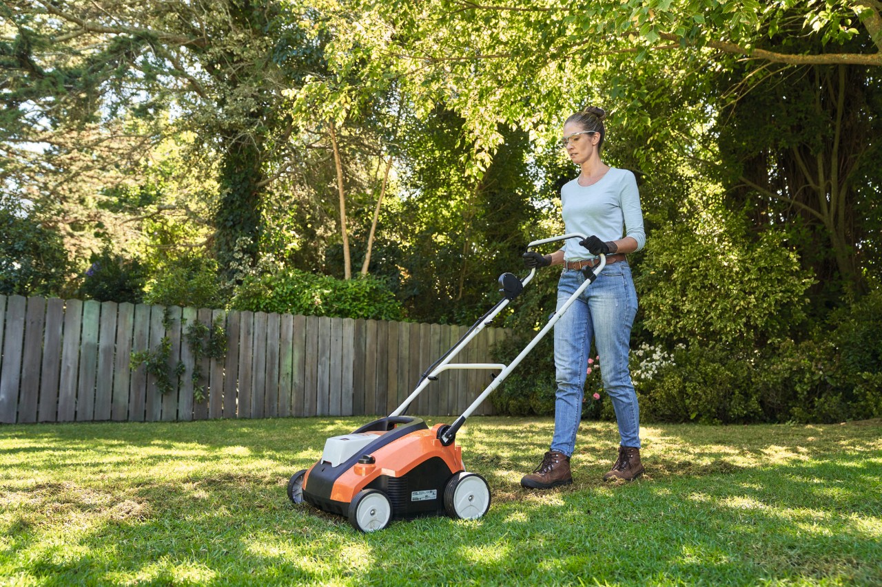 Uma mulher ventila o gramado no espaço verde com o arejador de grama a bateria STIHL RLA 240, equipada com óculos de proteção e luvas de trabalho.