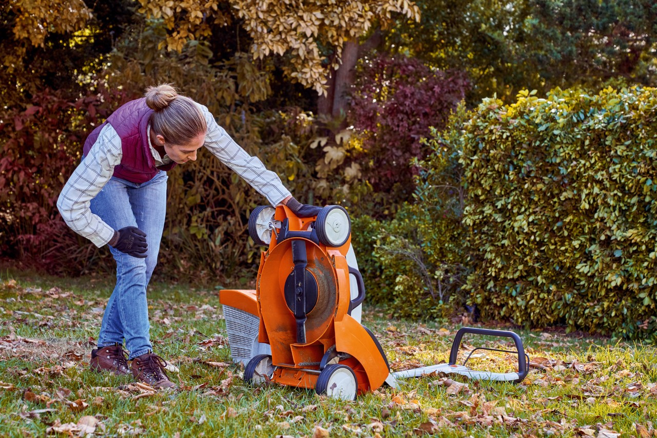 Mulher aplica os cuidados com a grama no outono ao inspecionar a parte inferior de um cortador de grama STIHL RMA 339 C 