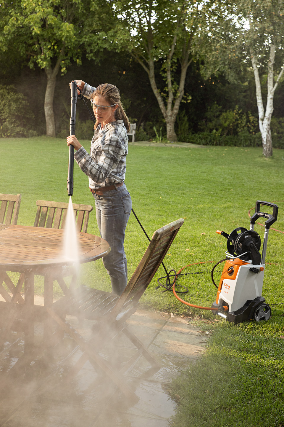 Uma mulher usa a lavadora de alta pressão STIHL RE 130 PLUS no exterior para limpar móveis de jardim