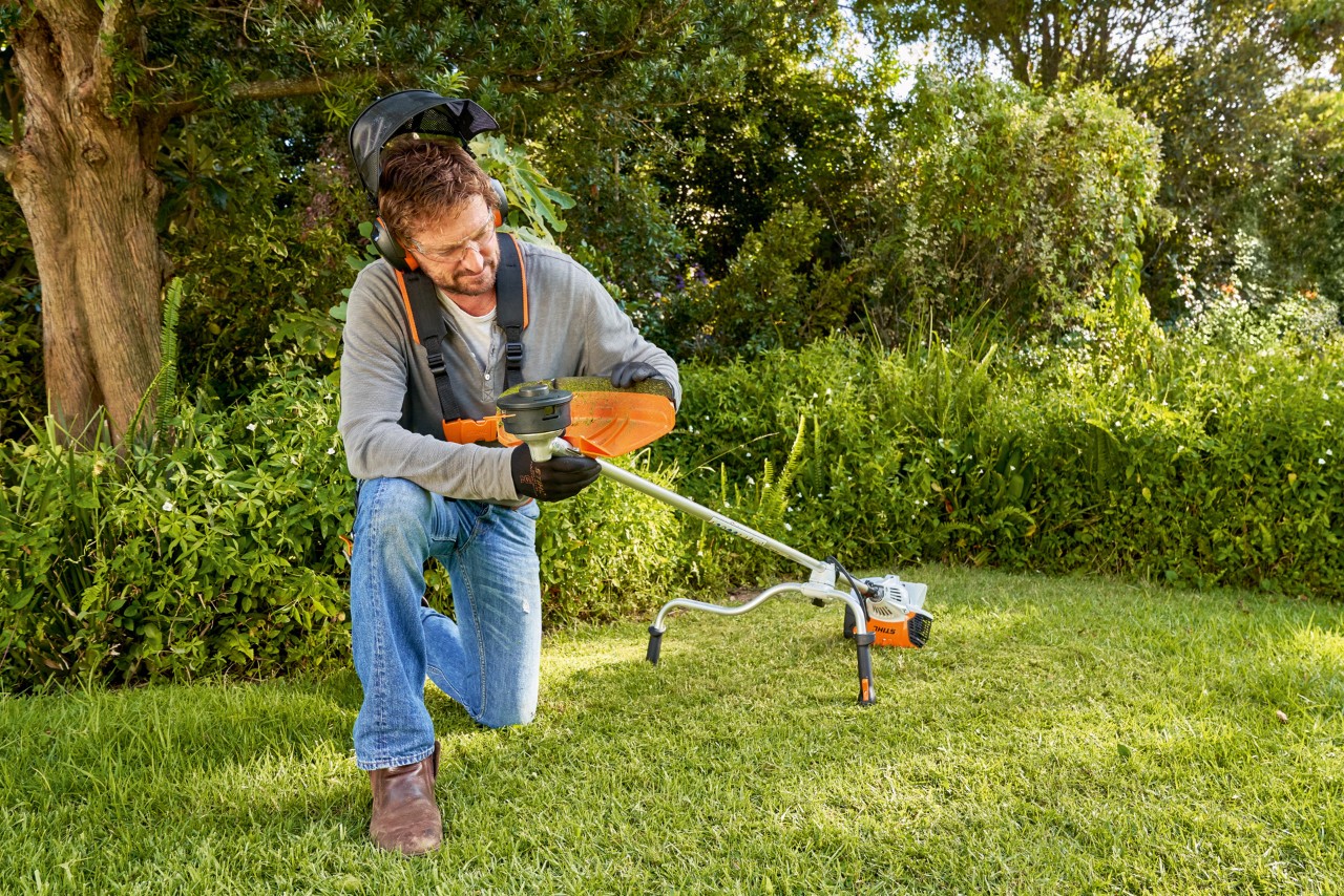 Um homem com equipamento de proteção demonstrando como substituir uma ferramenta de corte de uma roçadeira num jardim 