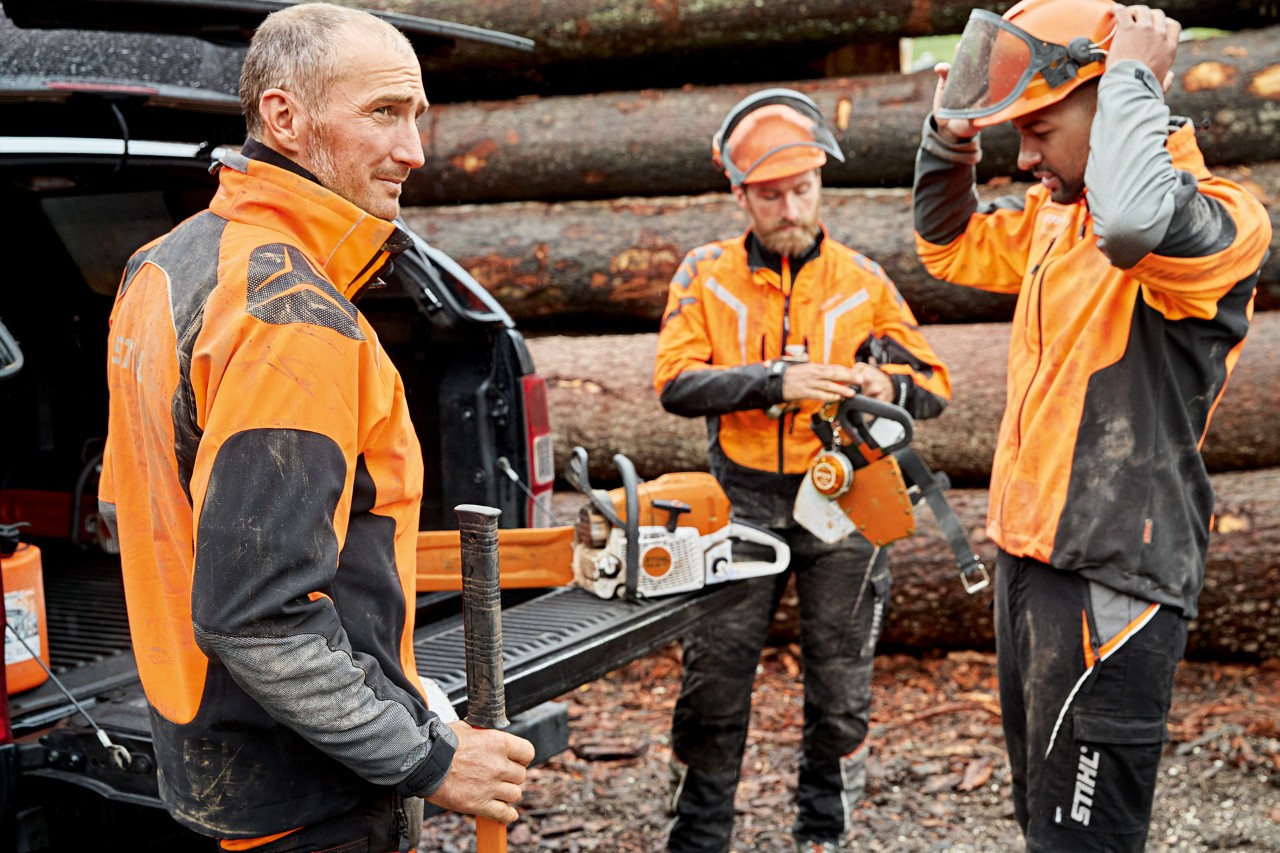Três homens com vestuário de proteção STIHL à frente de uma pilha de madeira a preparam-se para trabalhar com uma motosserra STIHL