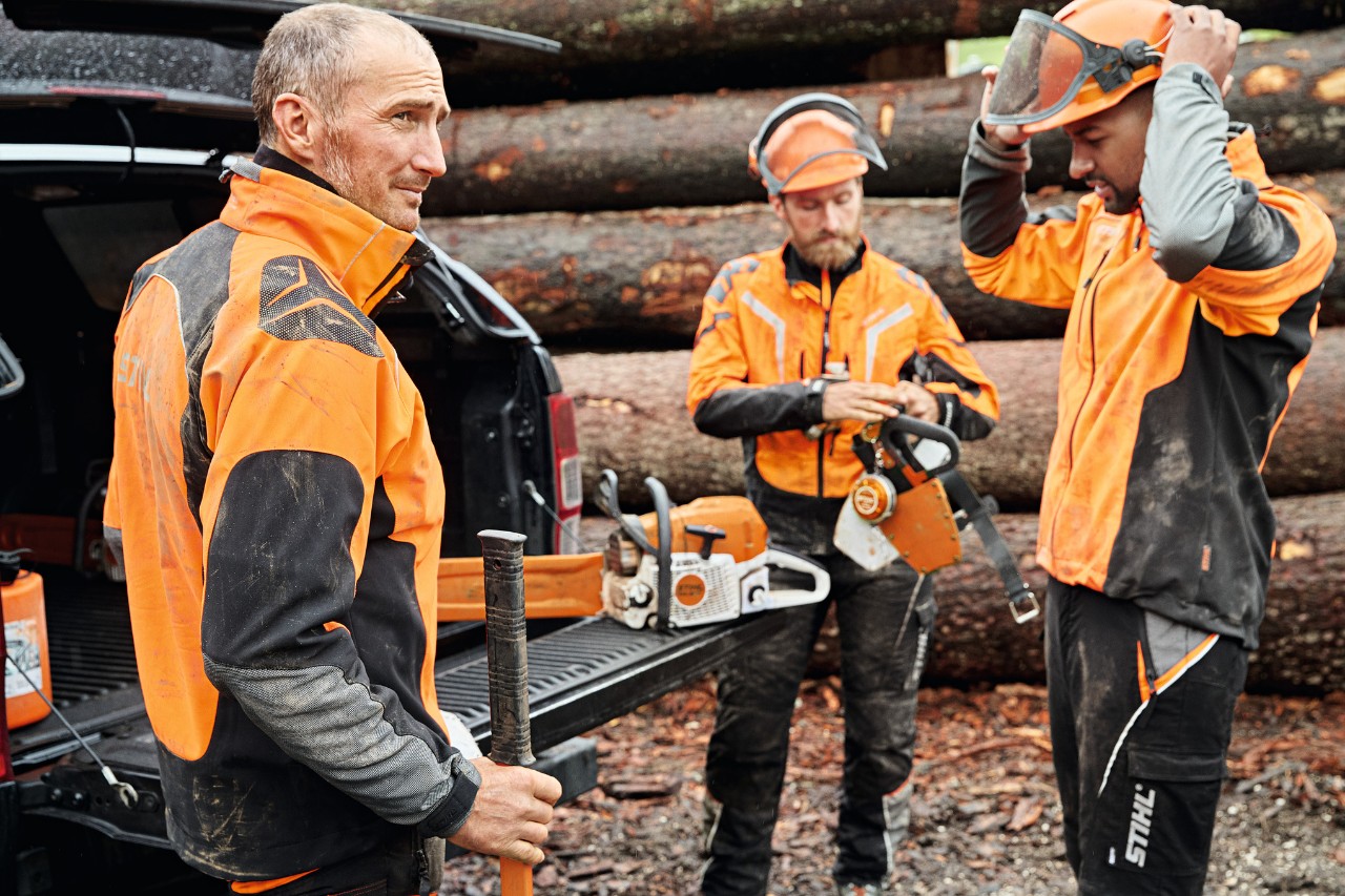 Três homens com equipamento de proteção individual STIHL e com máquinas a bateria STIHL junto à mala aberta de um veículo; em segundo plano, uma pilha de madeira