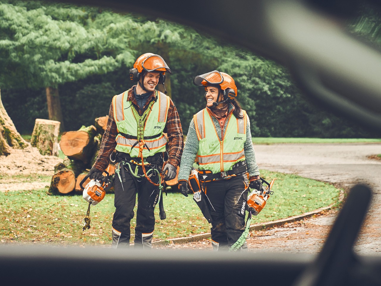 Um homem e uma mulher com motosserras sem fio STIHL e um podador sem fio STIHL caminham sorridentes em direção a um carro  