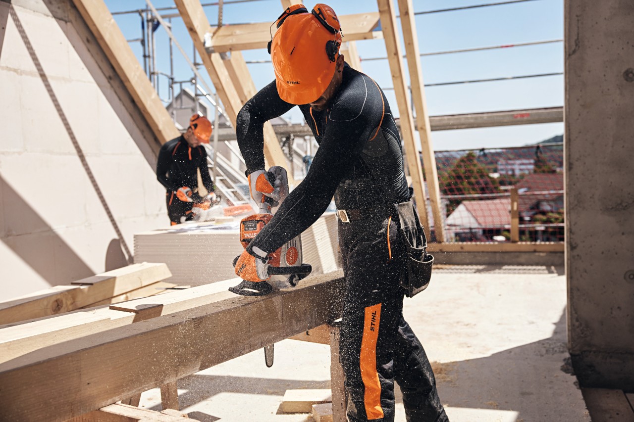 Um profissional da STIHL, numa estrutura de telhado, utiliza uma motosserra a bateria para cortar uma viga de madeira e, em segundo plano, outro trabalhador trabalha com uma motosserra a bateria